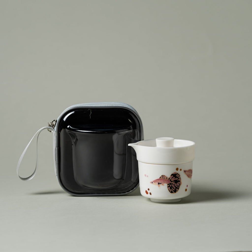 Lidded white porcelain brewer nested over guppy-pattern cup beside travel case