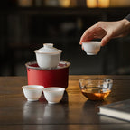 Hand lifting a white tasting cup beside a lidded gaiwan, two cups, clear pitcher and red travel case