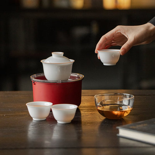 Hand lifting a white tasting cup beside a lidded gaiwan, two cups, clear pitcher and red travel case