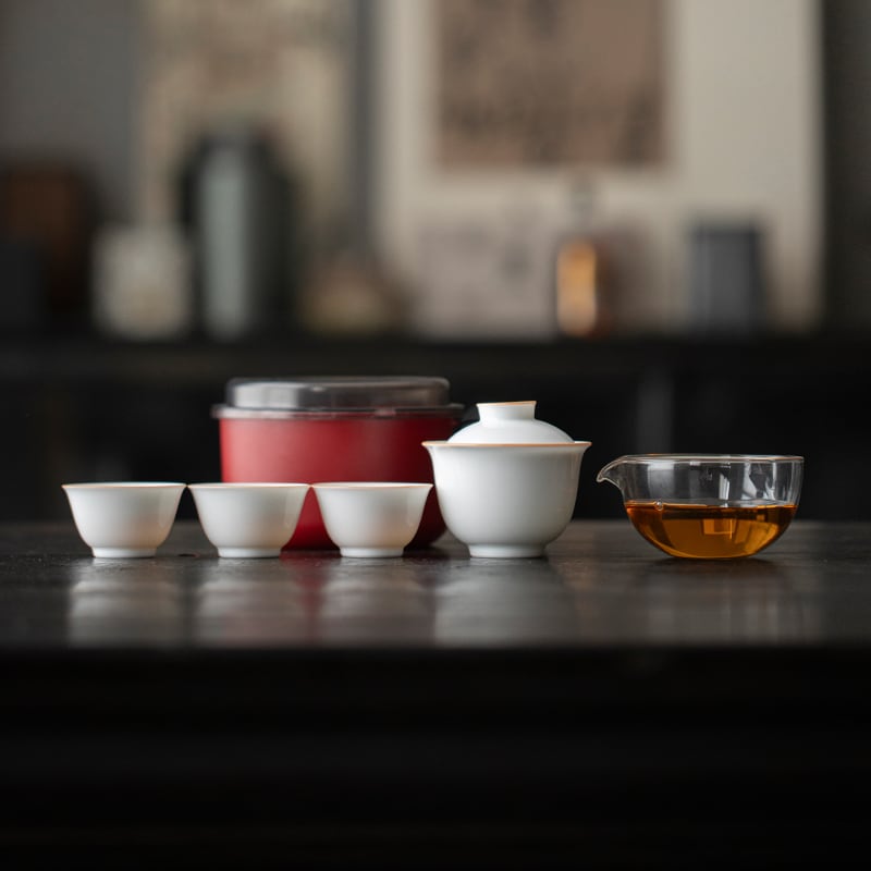 White gaiwan, three tasting cups and clear pitcher lined up beside a red round travel case