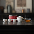 White gaiwan, three tasting cups and clear pitcher lined up beside a red round travel case