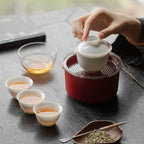 Hand lifting the lid of a white gaiwan with three cups, clear pitcher and red travel case on a dark table