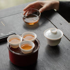Hand holding a clear pitcher above three white cups on a red travel case, with a gaiwan beside them