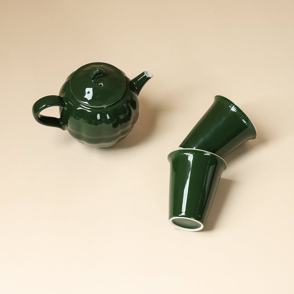 Overhead arrangement of a dark green round teapot and two tapered cups on a beige surface