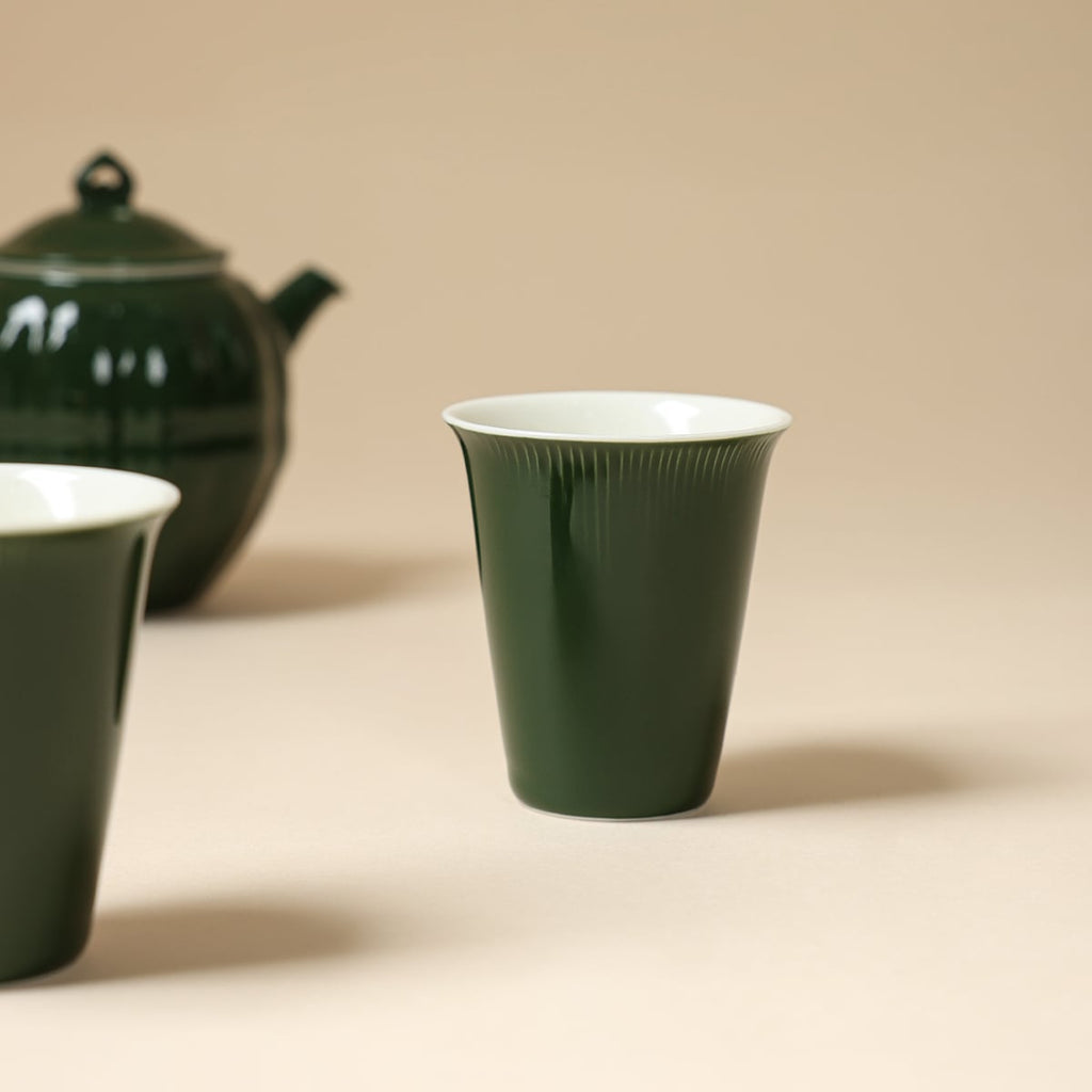 Dark green tapered cup with white interior, with the teapot and second cup softly blurred behind