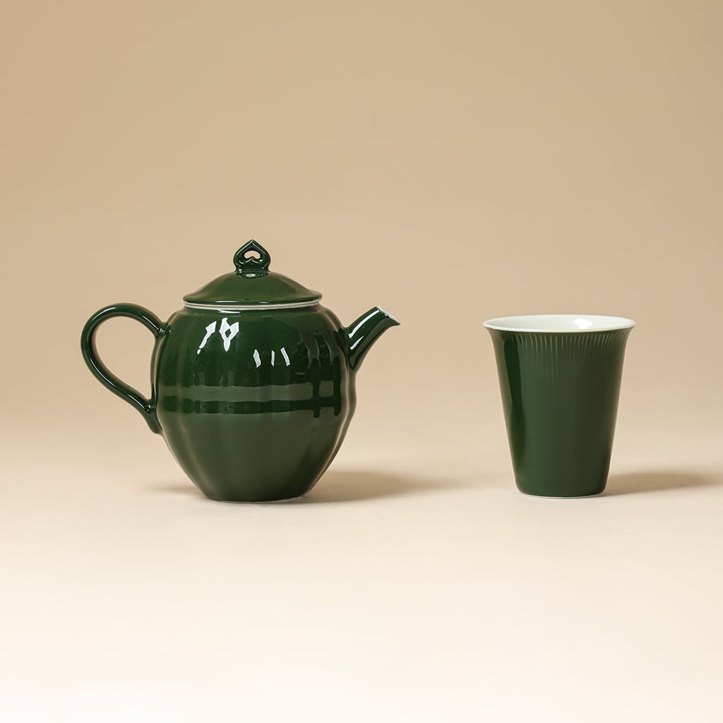 Dark green round teapot beside a matching tapered cup on a beige background
