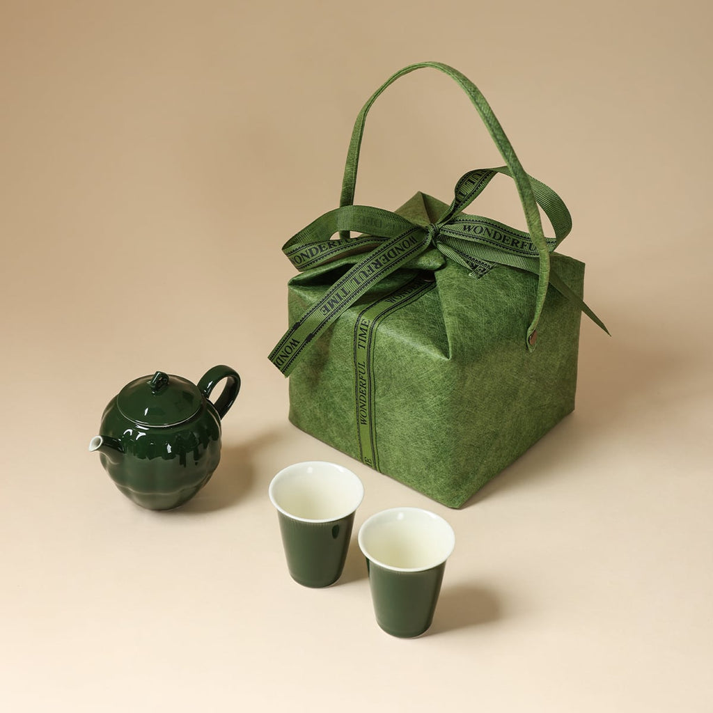 Dark green teapot and two cups arranged beside a square green carry bag