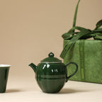 Side view of a dark green round teapot with a green carry bag and cup in the background
