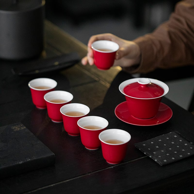 Hand holding a red-and-white tasting cup beside the gaiwan and remaining cups