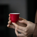 Hands holding the red-and-white gaiwan above a table set with matching tasting cups