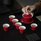 Hand lifting the white lid from the red gaiwan with matching tasting cups arranged nearby